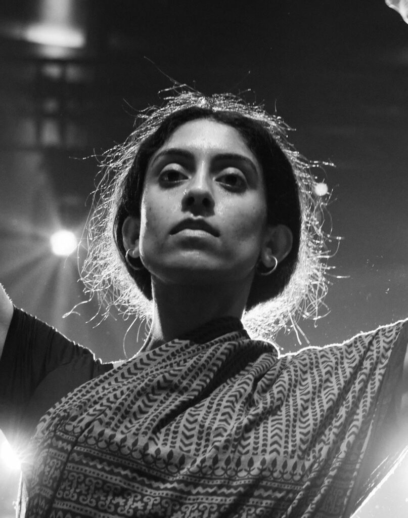 A black and white image with a black background with lights with Anjana, an Indian female, at the center of the photo. Anjana’s body is cropped, but hands are presumably stretched upwards in a circle. Her hair is loosely tied in the back in a low bun and she is wearing a black shirt with a multicolored dupatta.
