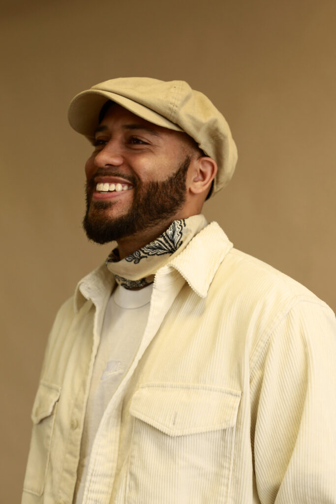 The image features a mixed-race man in his 30s, with short stylised facial hair. He is wearing a cream flat cap, collared shirt and necktie. He is smiling as he stands in front of a light brown wall.